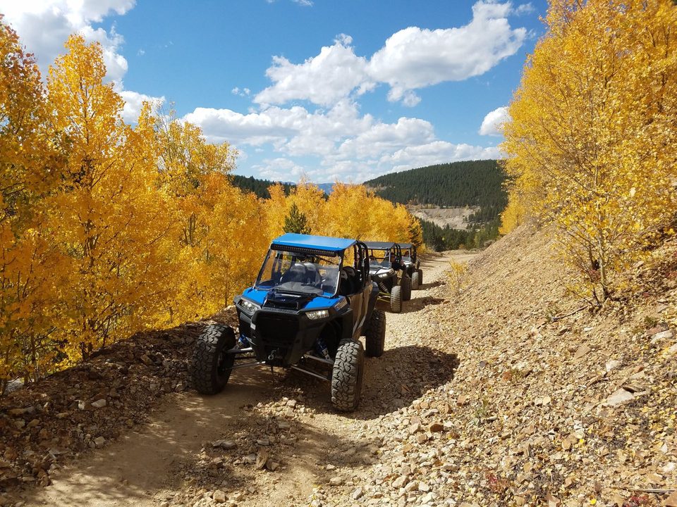 A blue Polaris RZR leading two more UTVs down a rocky mining road between gold aspens