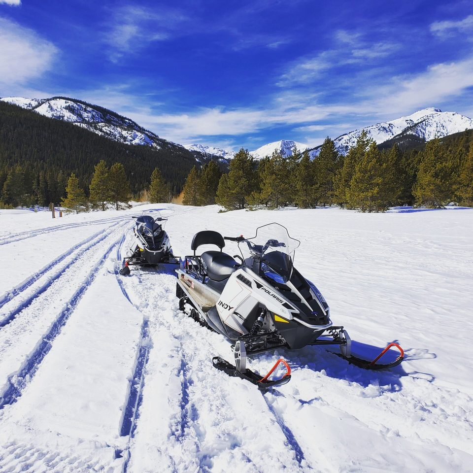 Two snowmobiles parked on a groomed trail under a deep blue sky, snowy peaks behind
