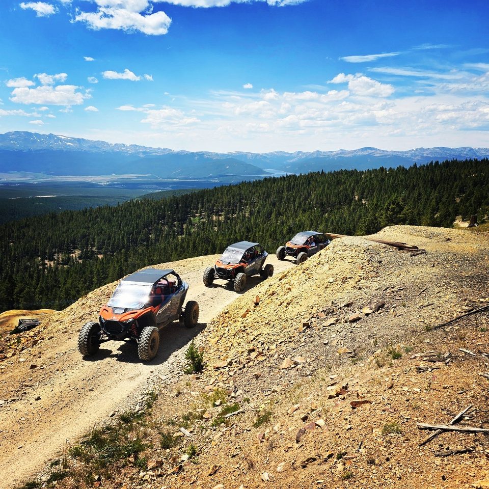 Three Polaris RZR side-by-sides climbing a narrow ridge road single file, a forested valley and distant snowy peaks below